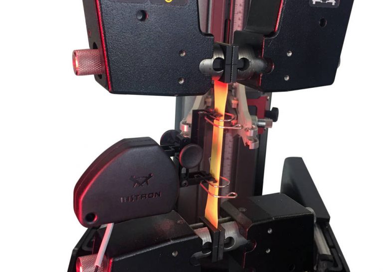 Instron testing system with grips performing tensile testing of a copper clad laminate strip specimen under red illumination