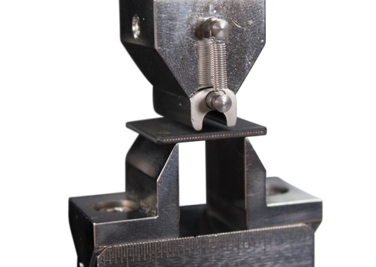 Close-up of Instron micro bend test fixture gripping a small electronic chip package specimen for three-point bend testing