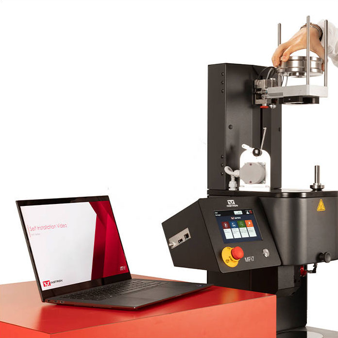 | Instron Macchina per prove sui materiali industriali con un utente che regola la morsa superiore mentre un laptop si trova su un tavolo rosso nelle vicinanze.