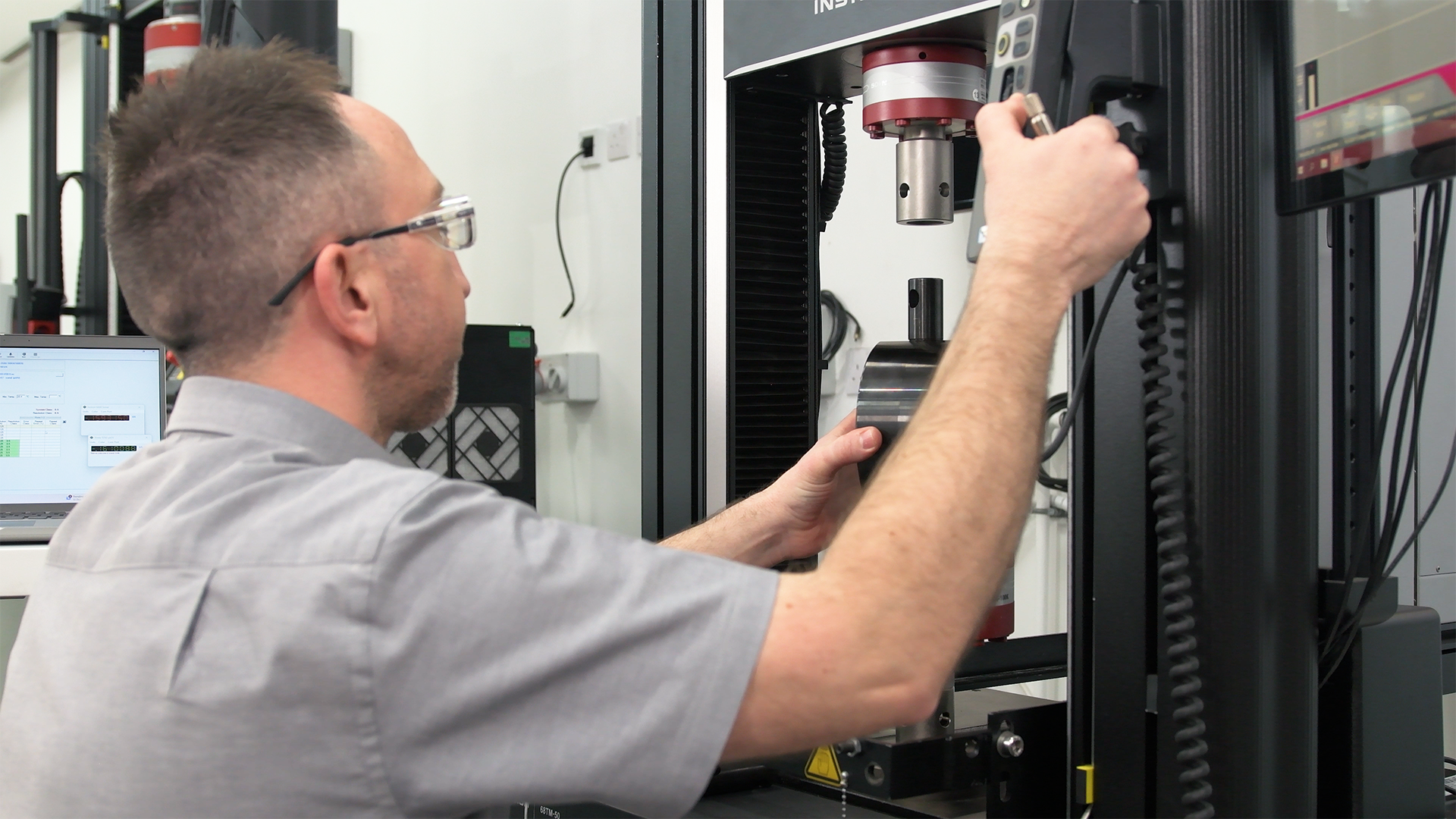 A technician operating an Instron universal testing machine, adjusting controls and positioning a cylindrical test specimen.