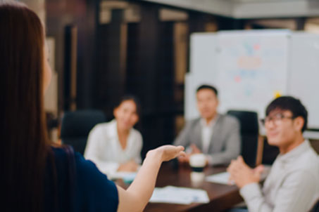 | Instron The back of a woman gesturing to a conference table with 3 other people