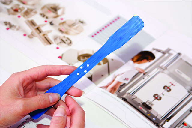 Hand holding a blue dogbone tensile test specimen alongside Instron training course materials