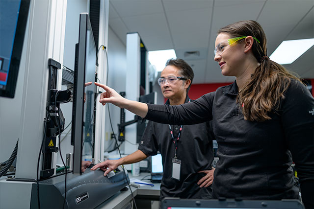 Student pointing at a touchscreen display on an Instron testing system with an instructor observing in the Norwood training facility