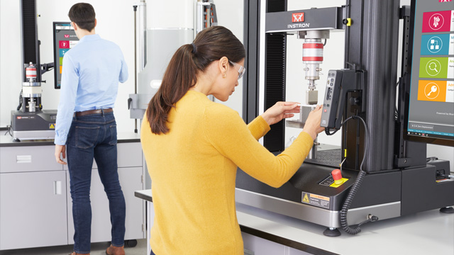| Instron A female lab technician in a yellow sweater and safety glasses operates an Instron 6800 Series universal testing machine with a touchscreen interface in a bright laboratory, while a male colleague works at a second Instron system in the background
