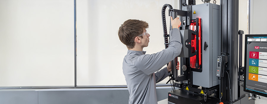 | Instron A male lab technician wearing safety glasses installs an Instron AVE3 Advanced Video Extensometer onto an environmental chamber mounted on a universal testing machine, with an operator dashboard with Bluehill Universal software visible to the right