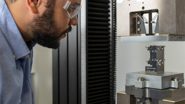 | Instron Engineer in safety glasses closely inspecting a composite test specimen loaded in an Instron compression testing fixture mounted between precision mechanical wedge grips.