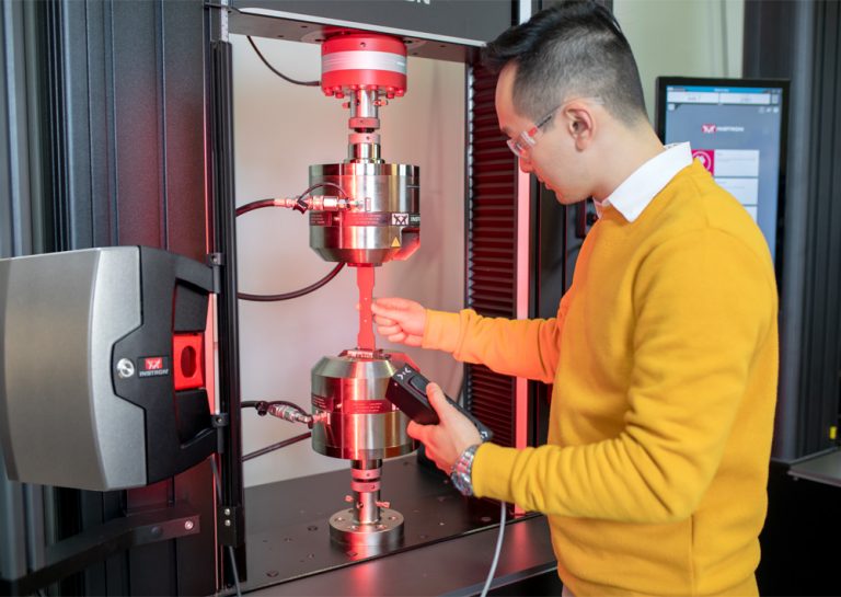 Lab operator in safety glasses and a yellow sweater loading a tensile specimen into hydraulic wedge grips on an Instron universal testing machine using a hydraulic wedge grip controller, with the AVE3 advanced video extensometer mounted on the left column illuminating the test space in red light