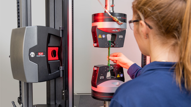 Operator loading a green plastic dogbone specimen into pneumatic side action tensile grips on a 6800 Series 68TM table model, with an AVE3 non-contact video extensometer positioned for strain measurement. | Instron