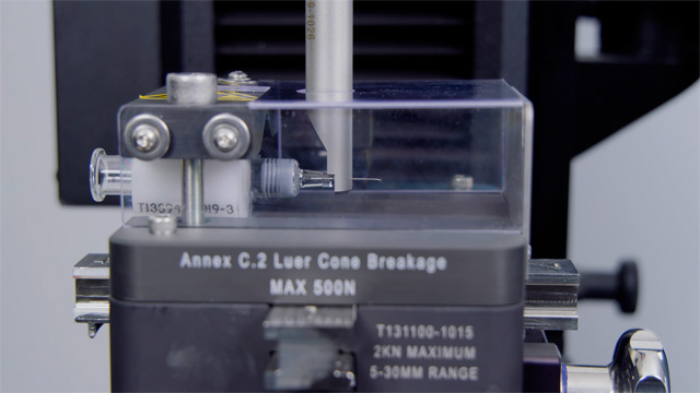 Annex C.2 Luer cone breakage fixture mounted on a 6800 Series testing system, used for syringe and Luer connector breakage testing in medical device quality control. | Instron