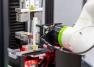 Close-up of a CT6 collaborative robot arm with green joint accent gripping and loading a white autoinjector specimen into the fixture of an Instron testing machine, with red illumination visible beneath the test platform