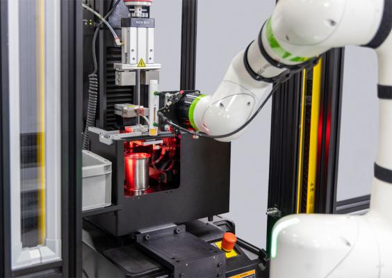 Wide-angle view of a CT6 collaborative robot arm extending into an Instron autoinjector testing machine to load a specimen, with yellow light curtain safety sensors, a discard bin, and an emergency stop button visible in the foreground